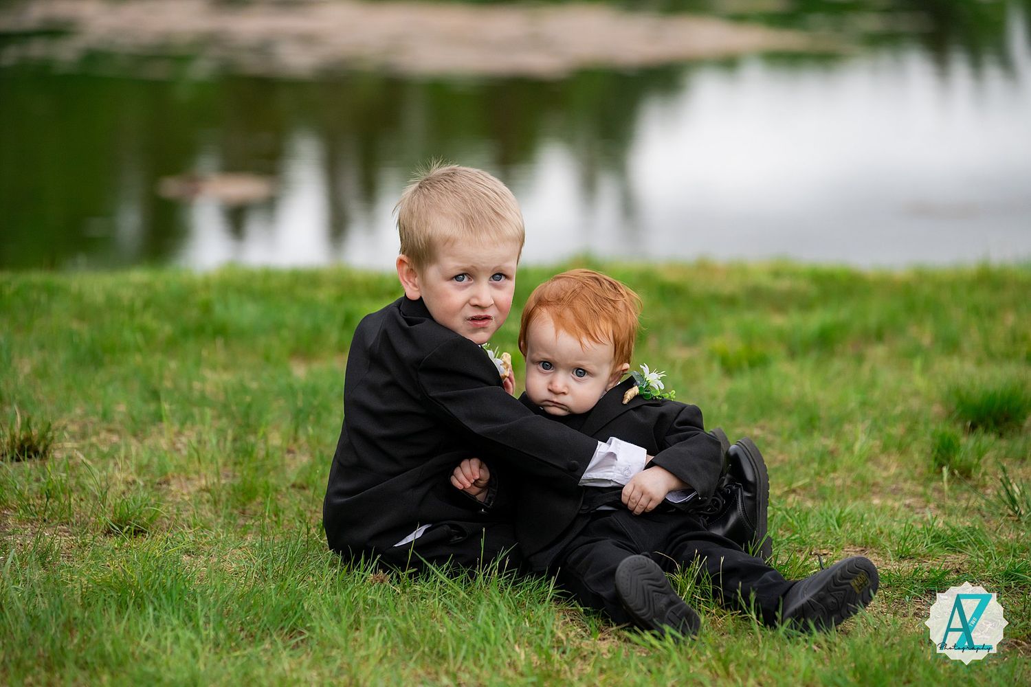 Two children in black clothing sit together on grassy ground near a pond.