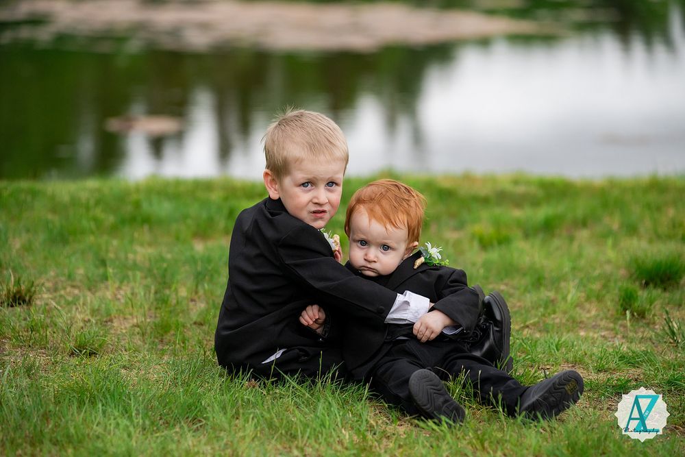 Two children in black clothing sit together on grassy ground near a pond.