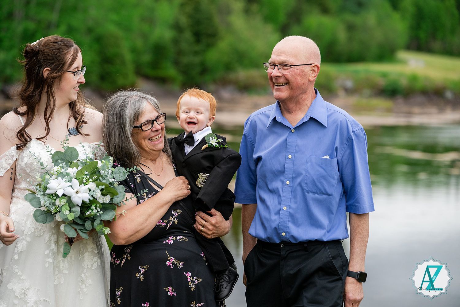 A joyful wedding party laughs together by a serene lake with a bride holding a white and green bouquet.