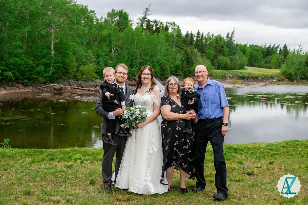Wedding party poses outdoors by a scenic pond with green trees and grass along the shoreline.