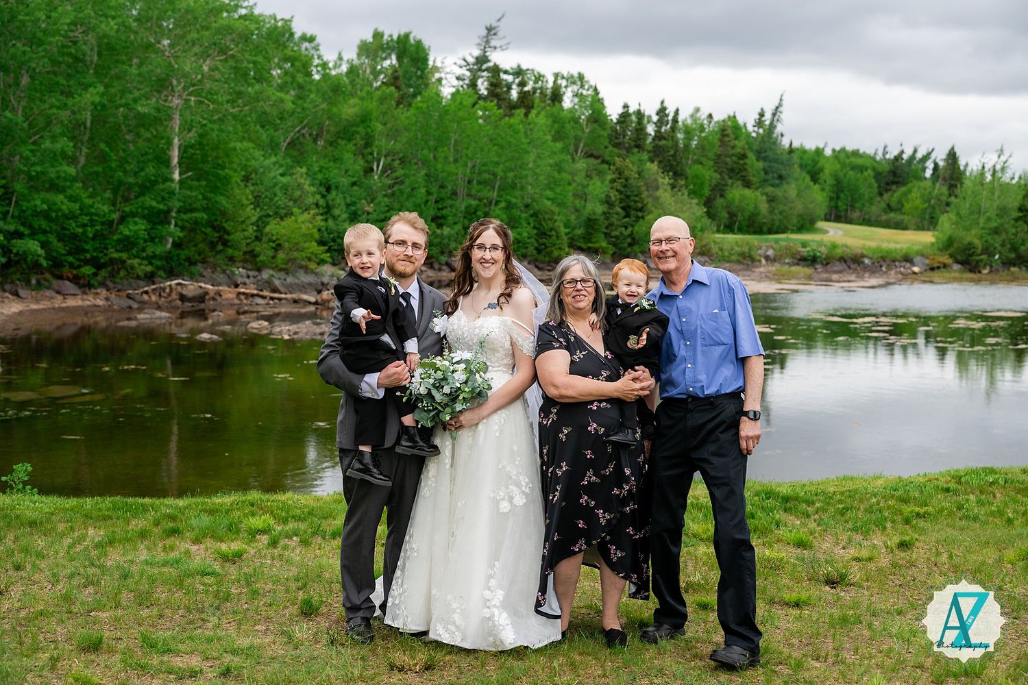 Wedding party poses outdoors by a scenic pond with green trees and grass along the shoreline.