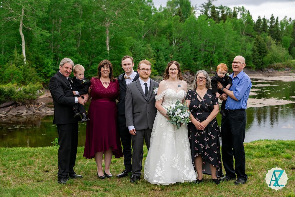 A wedding party poses against a beautiful lakeside backdrop with lush green trees and natural scenery.
