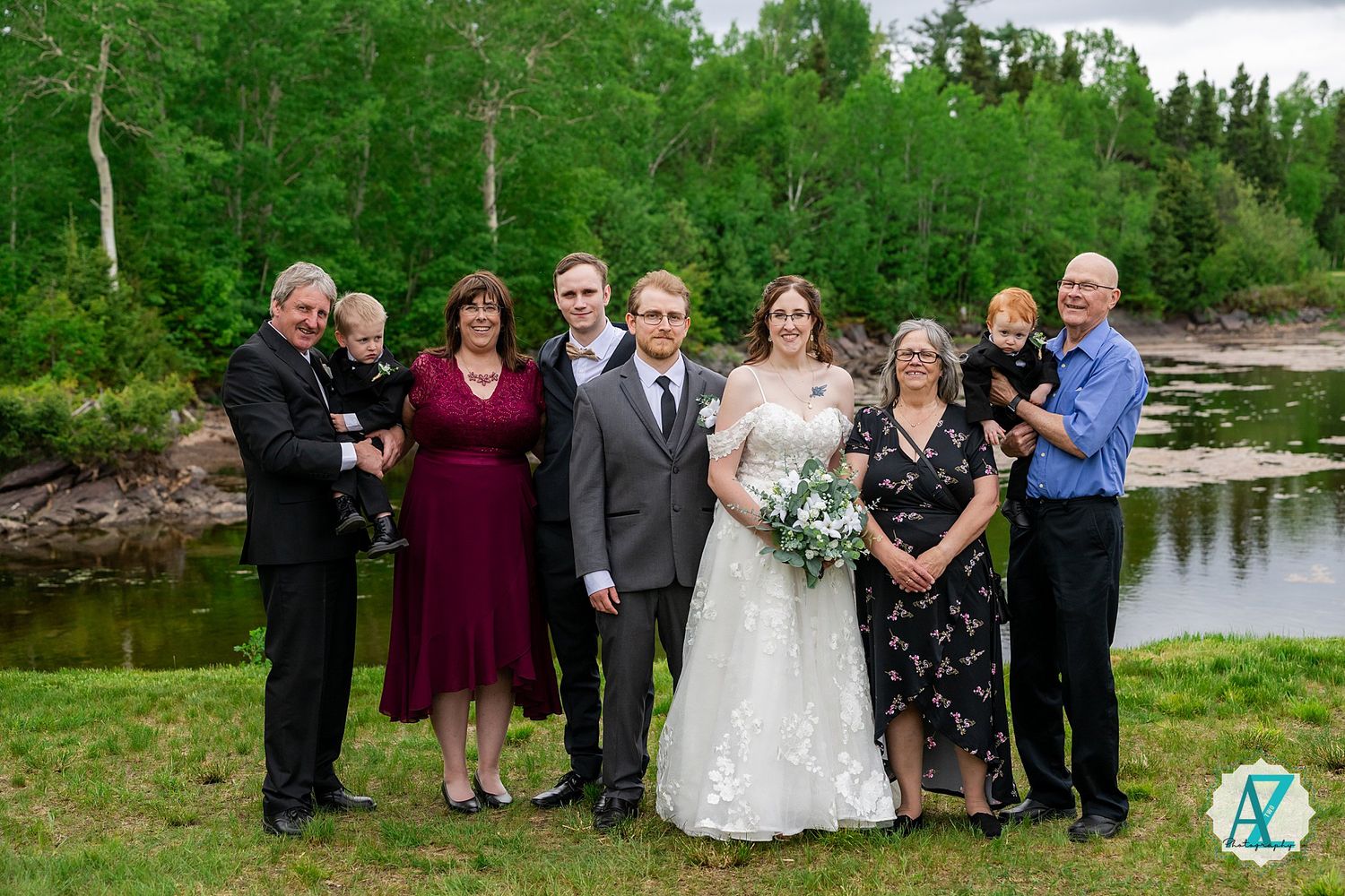 A wedding party poses against a beautiful lakeside backdrop with lush green trees and natural scenery.
