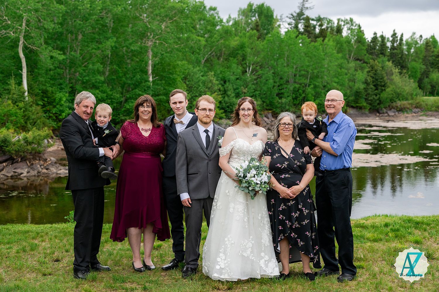 Wedding party poses together in a scenic outdoor setting by a tranquil lake with lush greenery in the background.