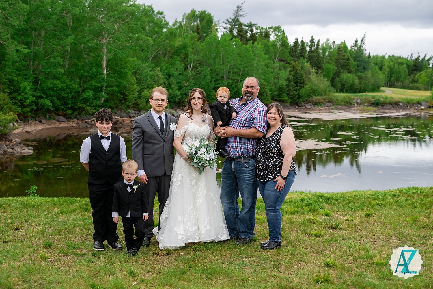Wedding party poses outdoors near a serene lake with green trees and natural landscape in the background.