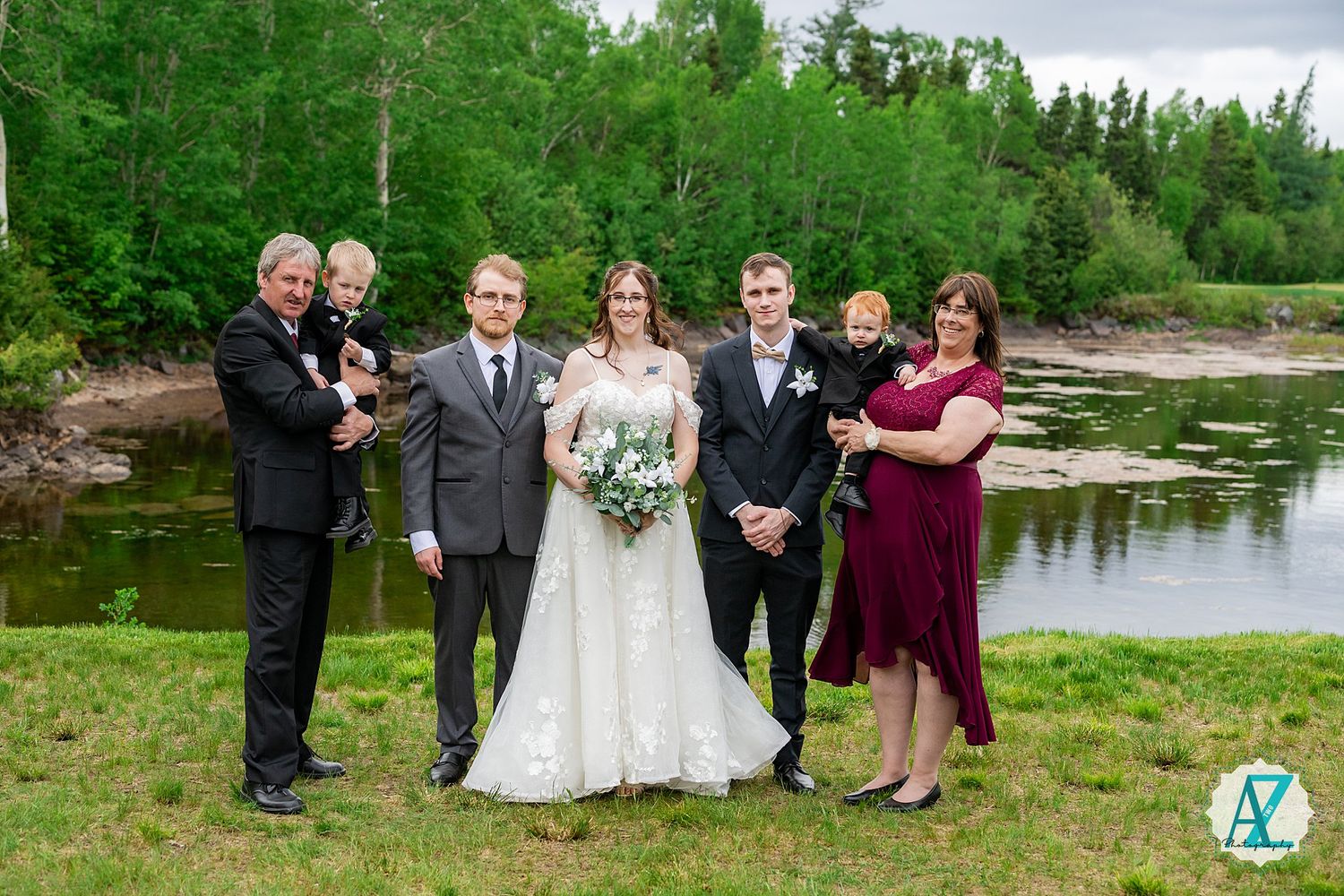 Wedding party poses beside a serene lake with lush green forest backdrop on a cloudy day.