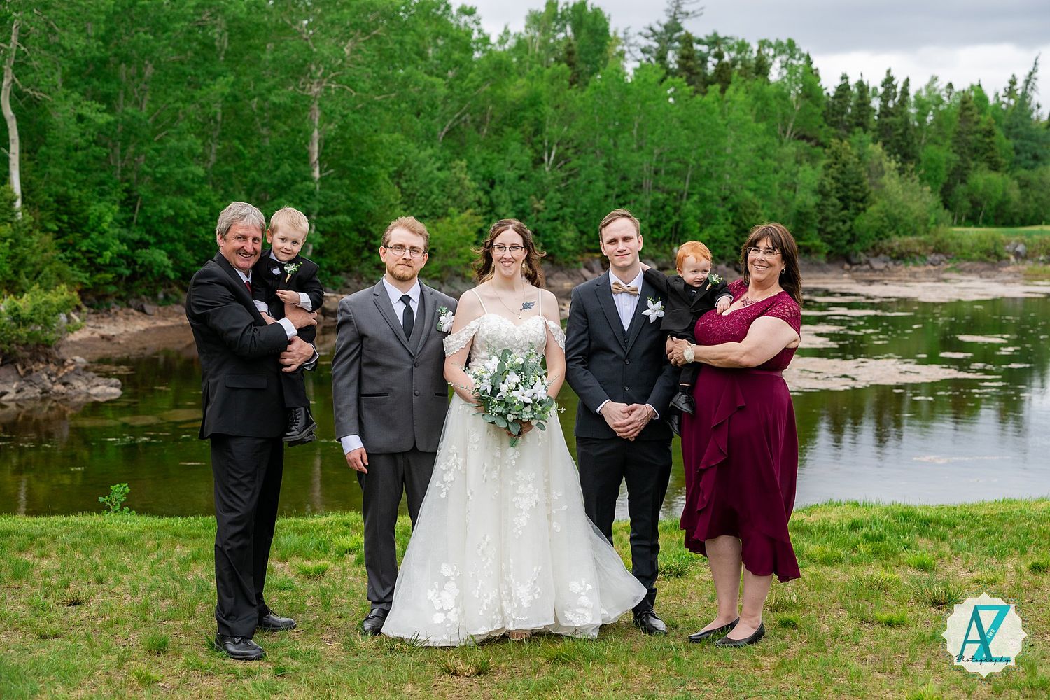 Wedding party poses together on grassy bank beside scenic lake with green forest in background.