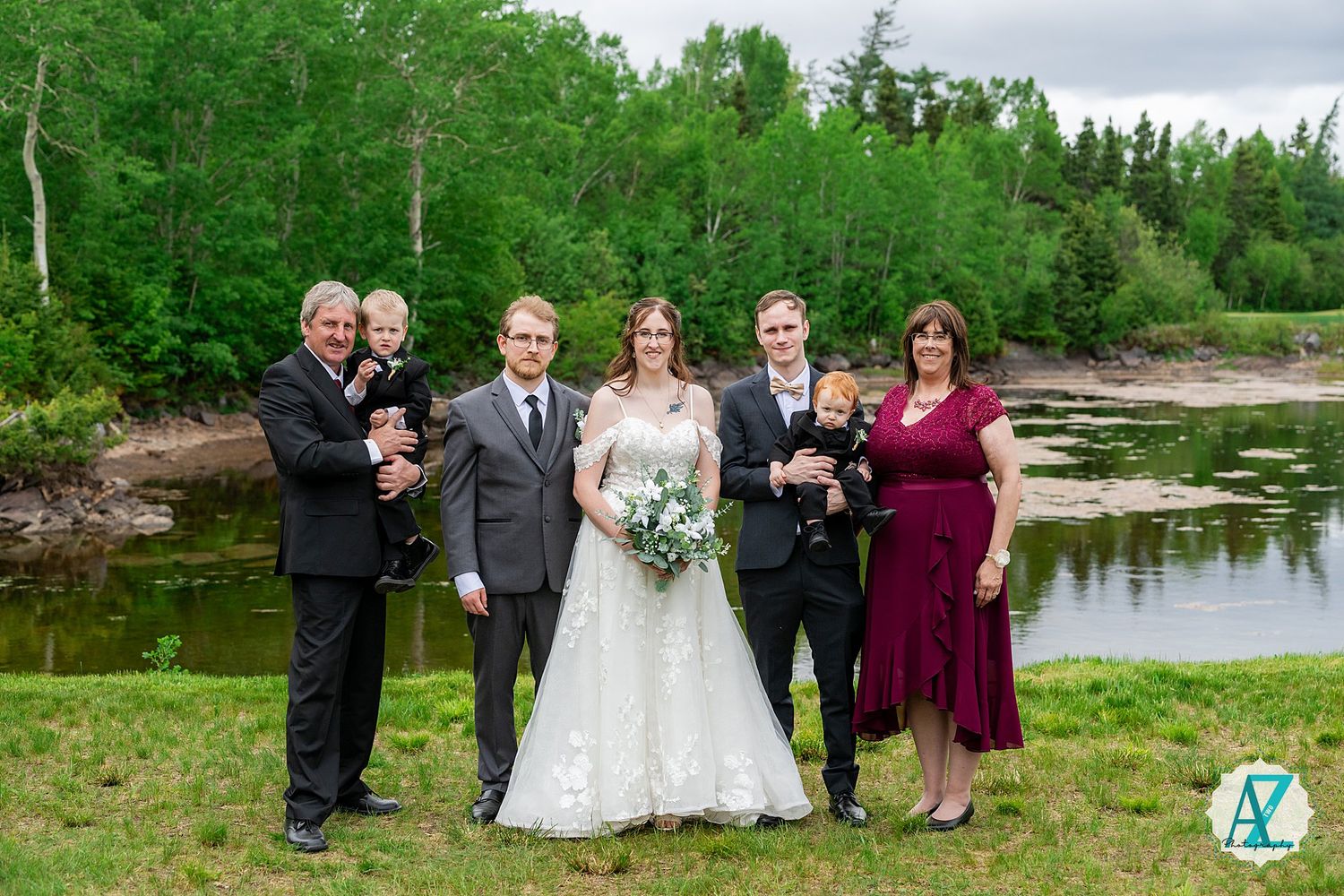 Wedding party poses for outdoor photos near a serene lake with lush green trees and natural scenery in the background.