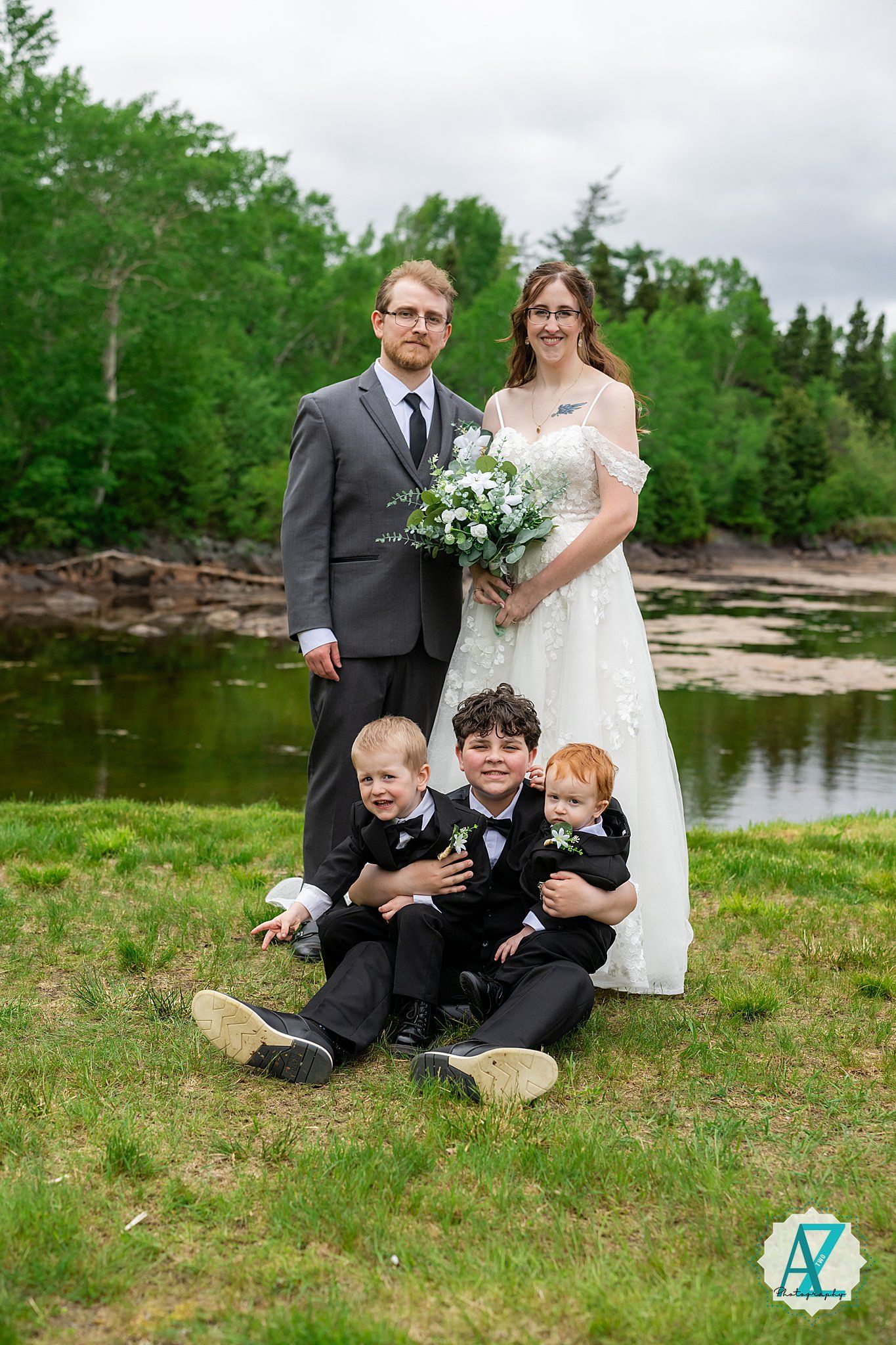 Wedding couple and three children pose for a portrait by a serene pond with trees in the background.