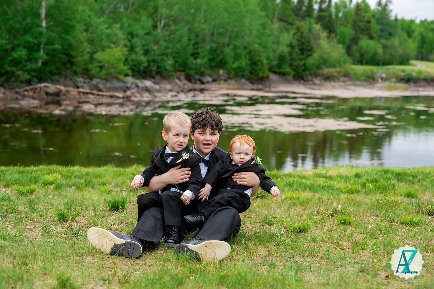 Three people sitting together on grass by a scenic lake with trees and rocky shoreline in the background.