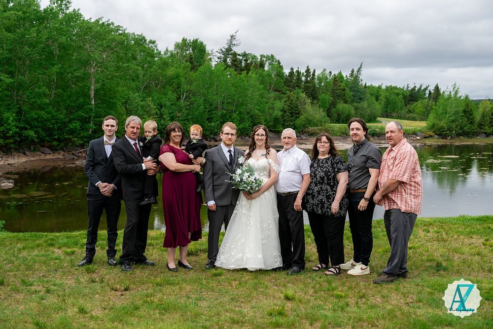 Wedding party members stand together for outdoor group photos by a serene lake on an overcast day.
