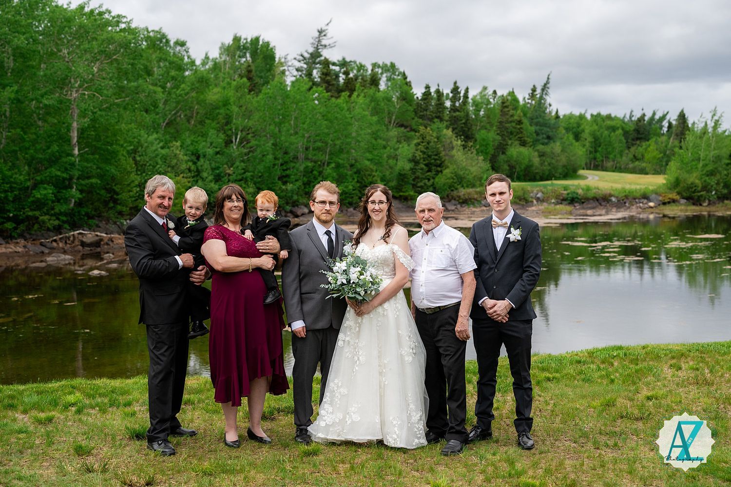 Wedding party gathered on green grass beside a tranquil pond with lush forest backdrop on cloudy day.