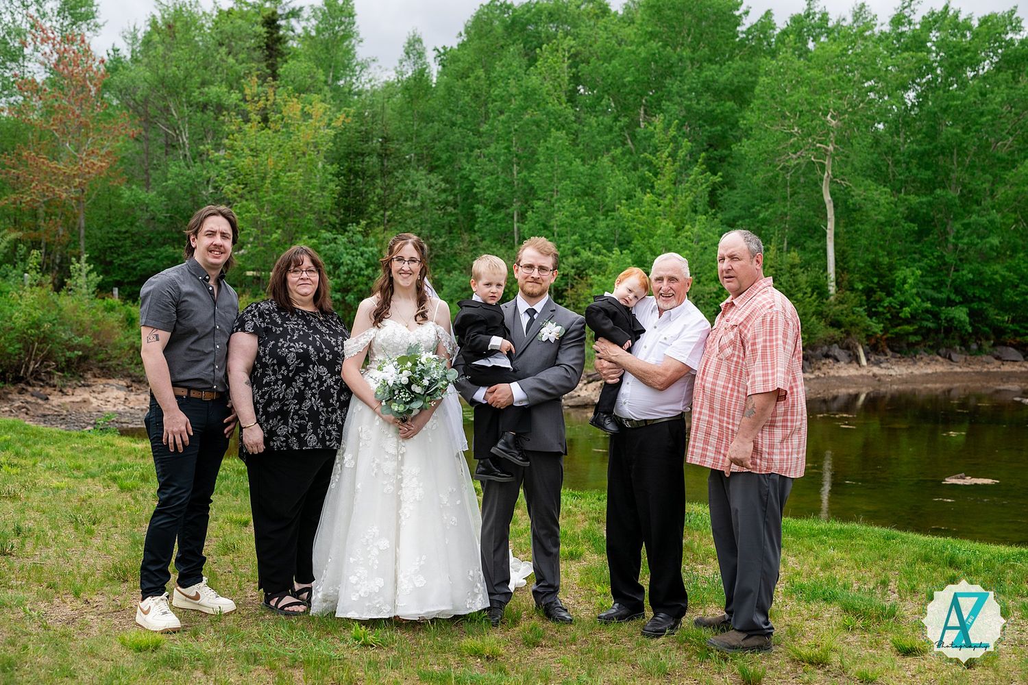 A wedding group poses together outdoors by a serene pond with lush green trees in the background.