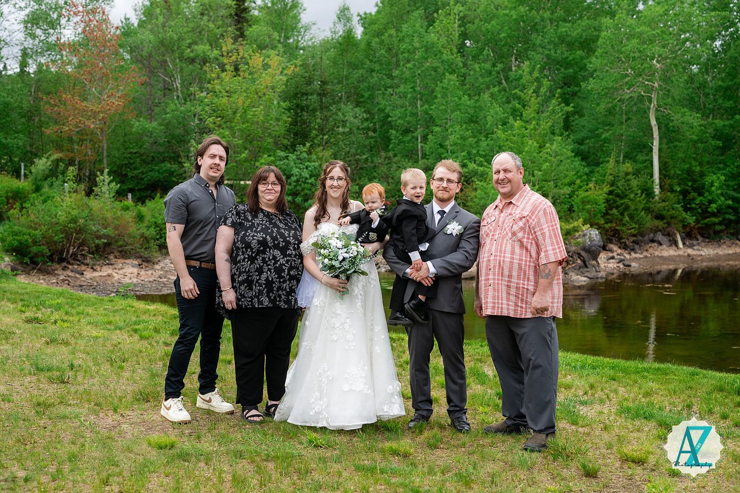 Wedding party poses together in scenic woodland setting surrounded by lush green trees and grass in summer.