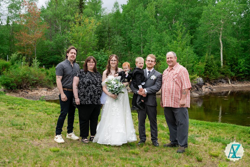 Wedding party poses together in scenic woodland setting surrounded by lush green trees and grass in summer.