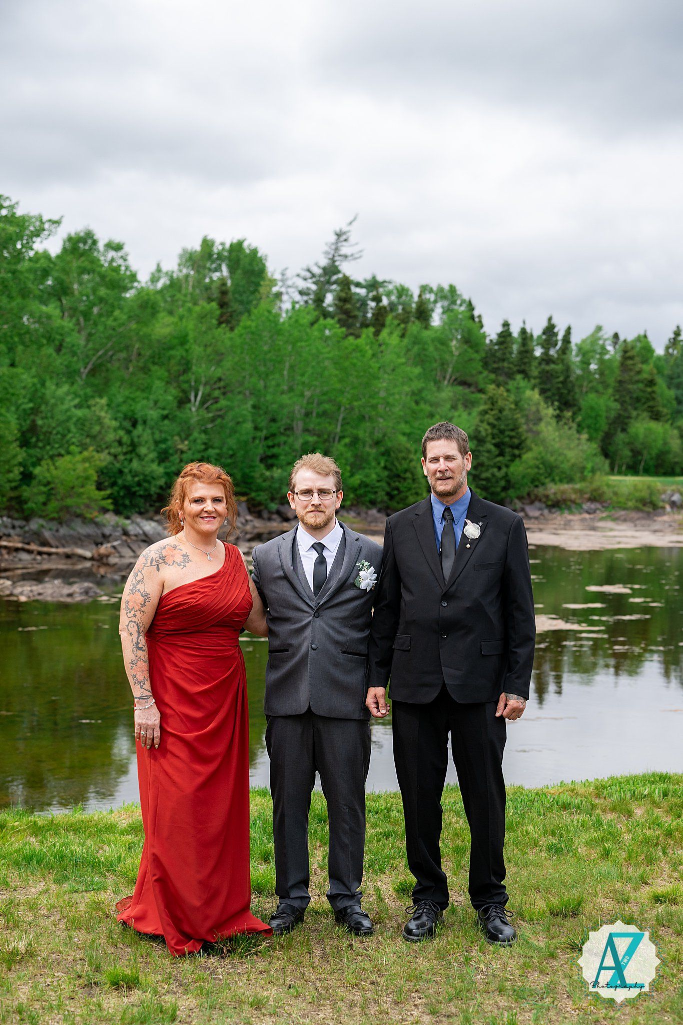 Wedding party in formal attire poses together by a lakeside on cloudy day with trees in background.