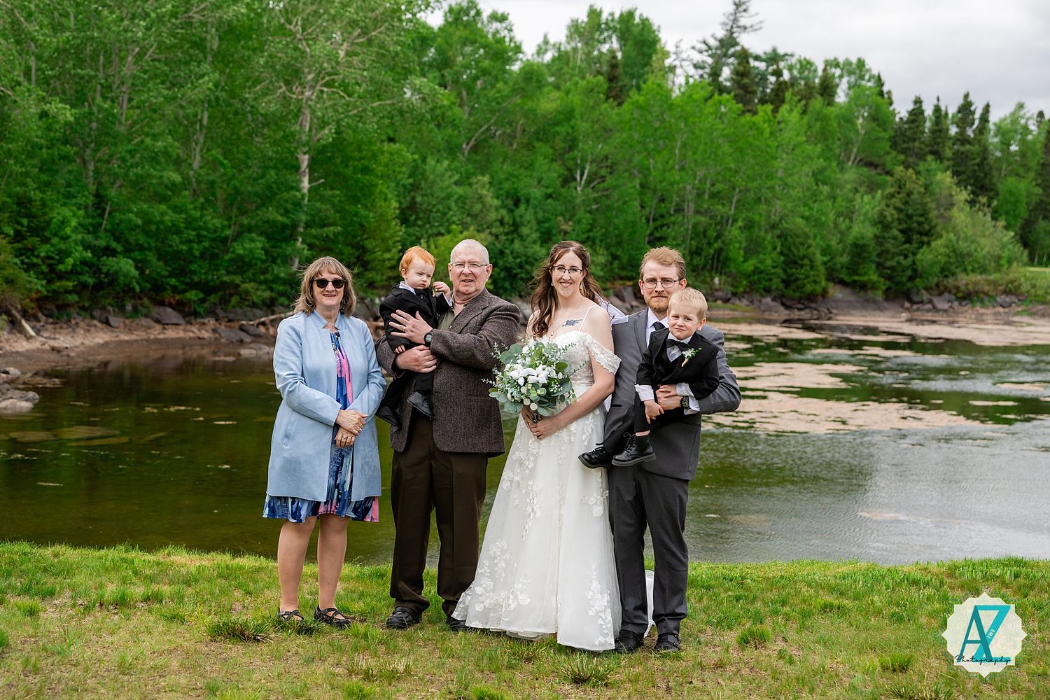 A small wedding party poses together on the grassy banks of a river with lush green forest in the background.