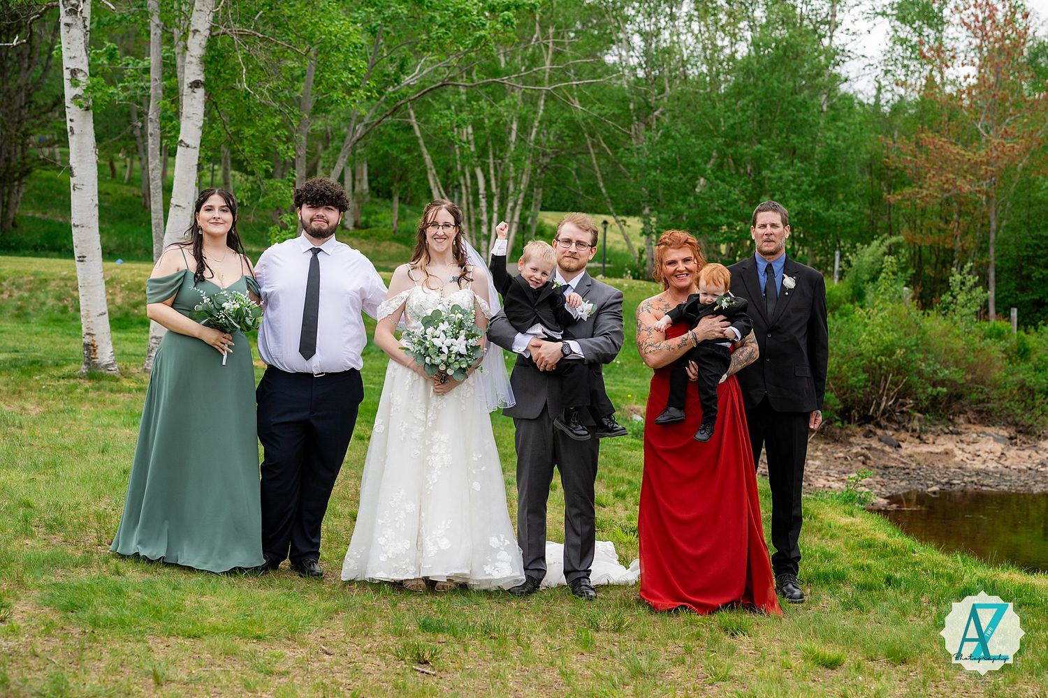 Wedding party stands in a line outdoors with birch trees and a pond in the background wearing formal attire.