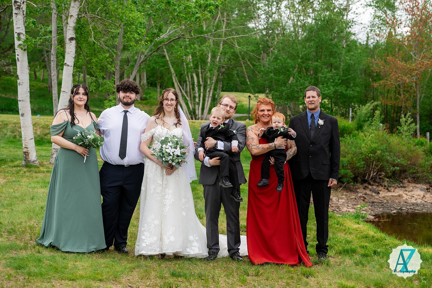Wedding party in formal attire poses together outdoors with birch trees and greenery in the background.
