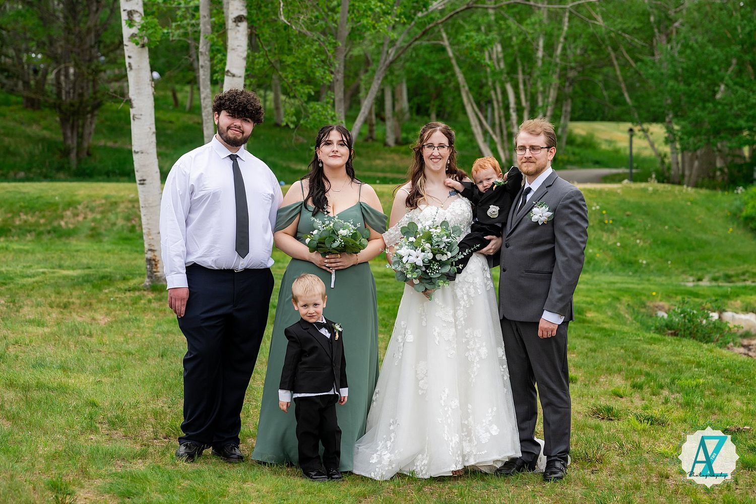 Group of five people dressed formally, including a bride in white lace gown, posing together outdoors in wooded setting.