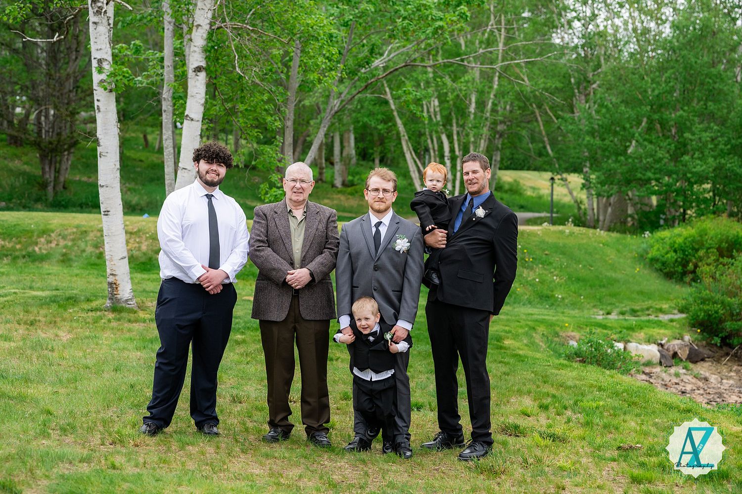 A wedding party poses outdoors in a green garden setting surrounded by trees and natural landscaping.
