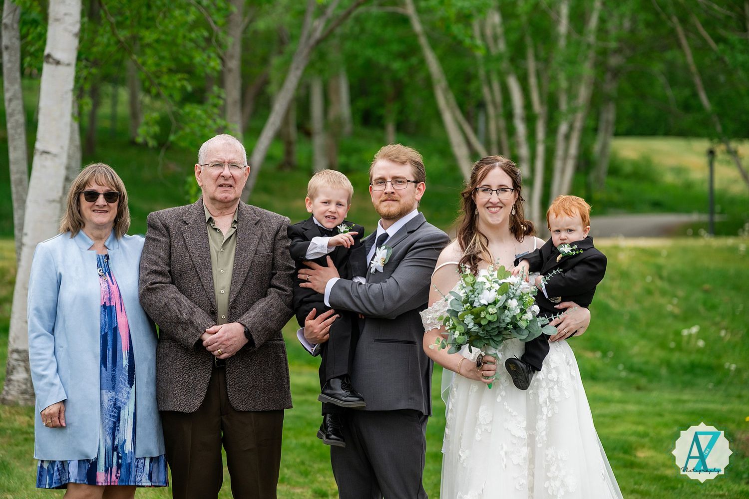 Wedding party stands together outdoors on a sunny spring day with trees and greenery in background.