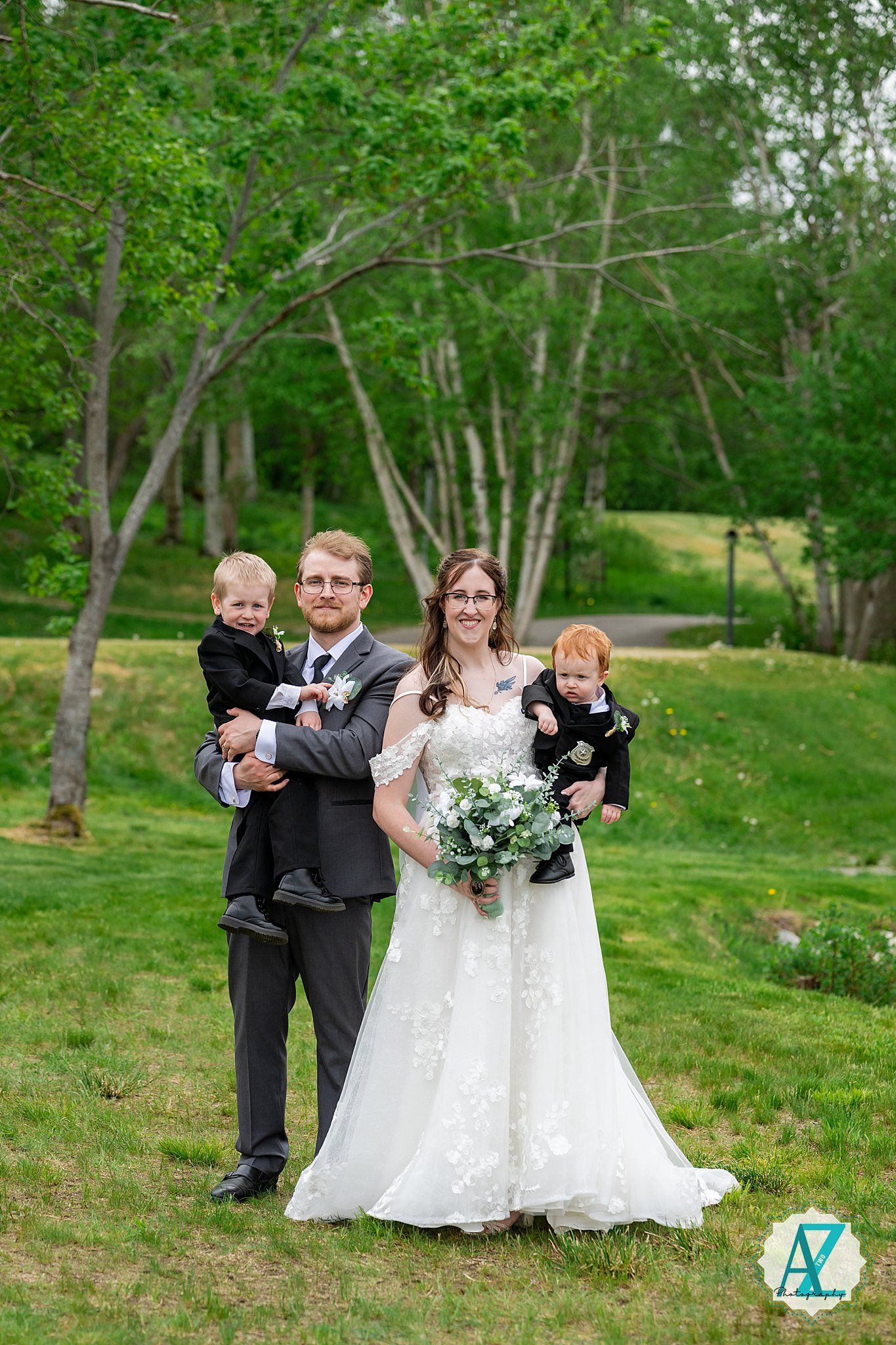 A wedding couple stands together outdoors on a grassy slope surrounded by green trees and foliage.