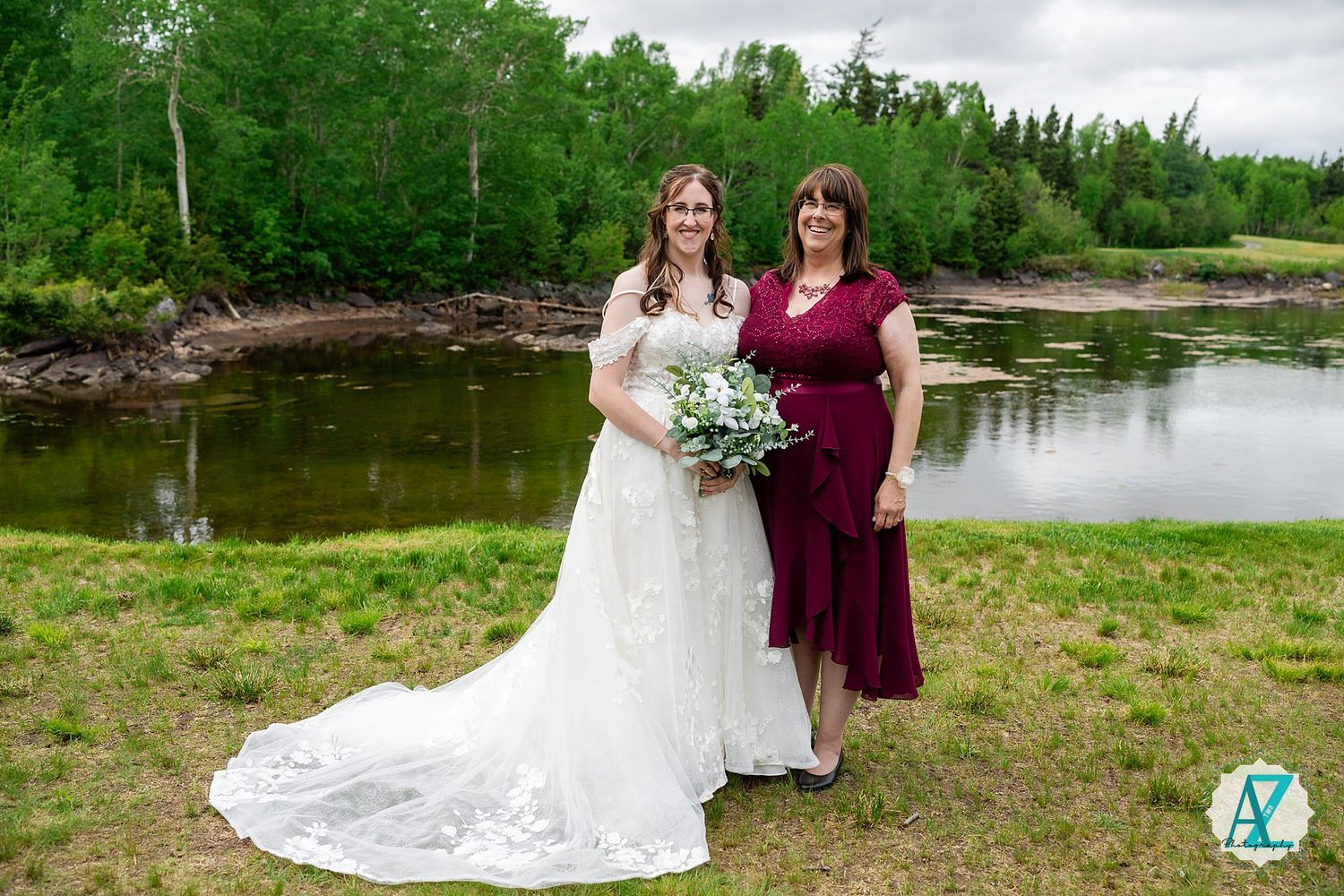 Two people in formal attire stand at the edge of a scenic pond with trees and greenery in the background.