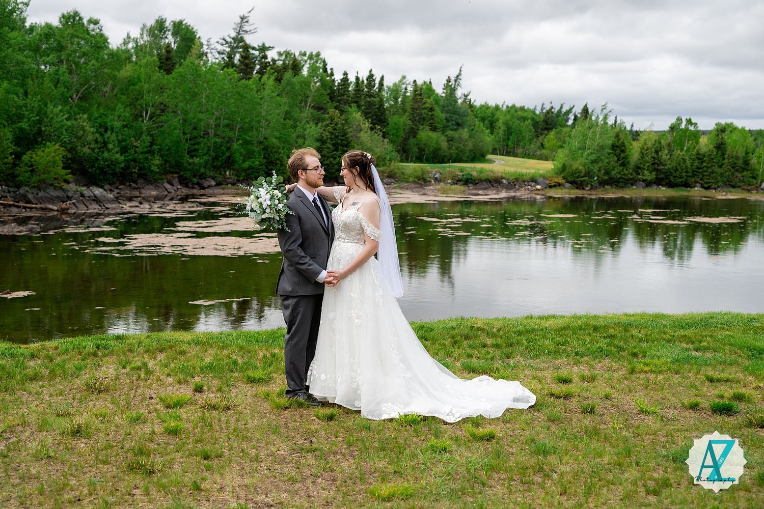 A couple in formal wedding attire stands on the grassy shore of a serene pond surrounded by lush forest.
