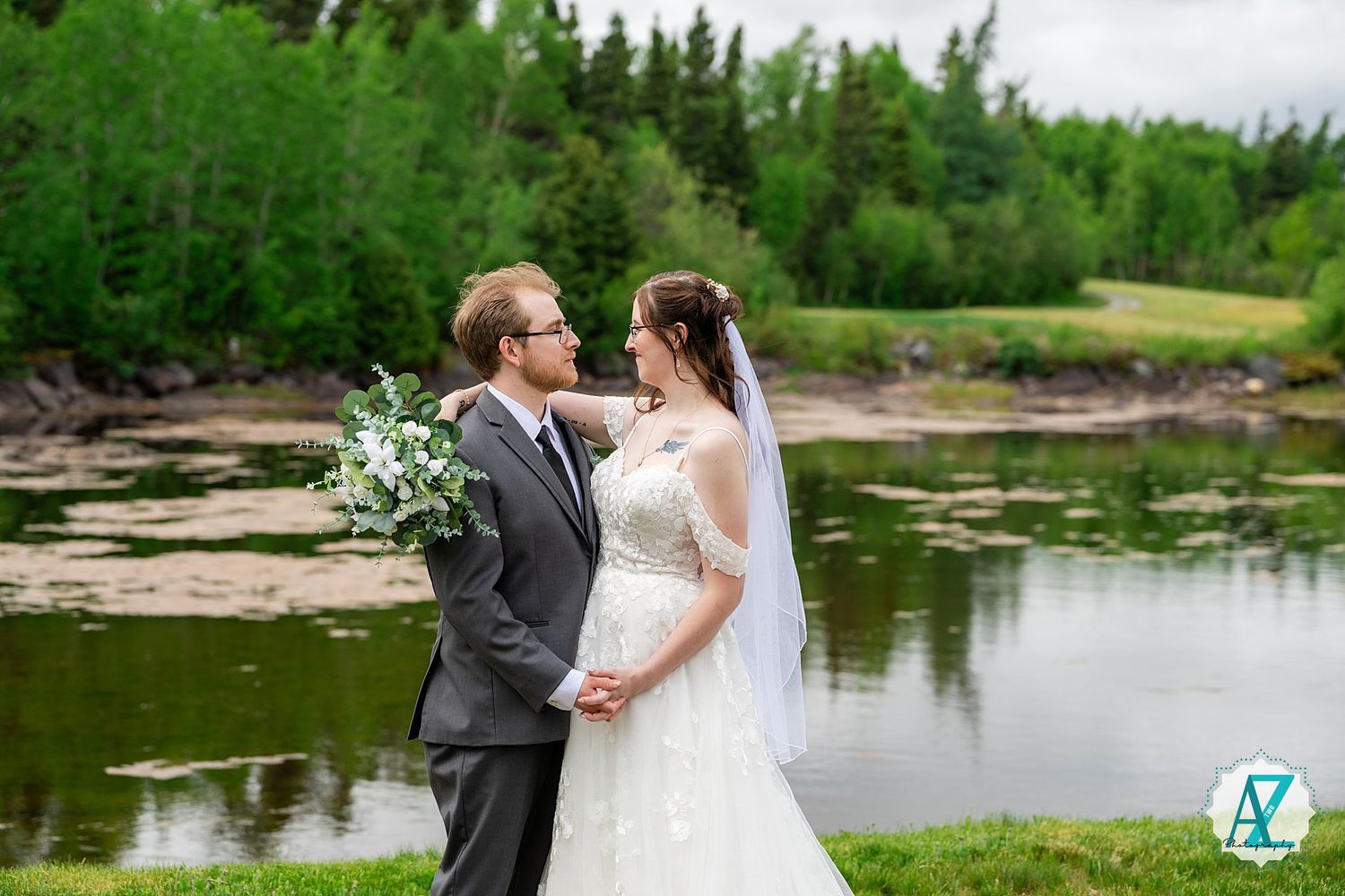 Bride and Groom share a tender moment while standing near a serene lake with green forest backdrop at their wedding.