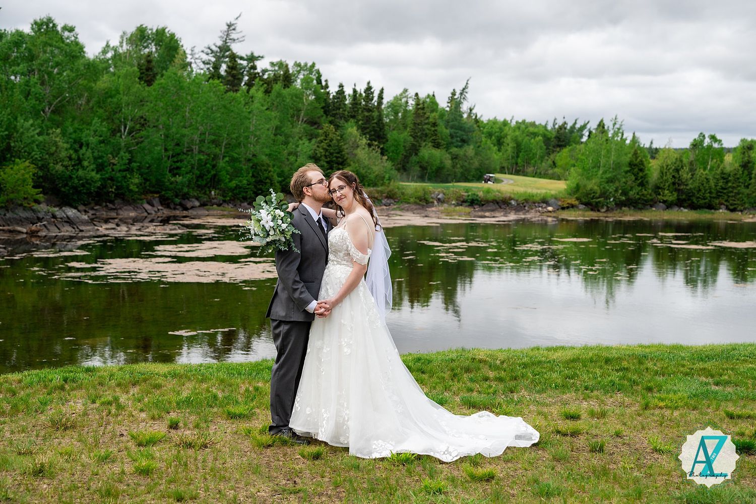 A romantic couple embraces by a serene pond surrounded by lush green trees and grass under an overcast sky.