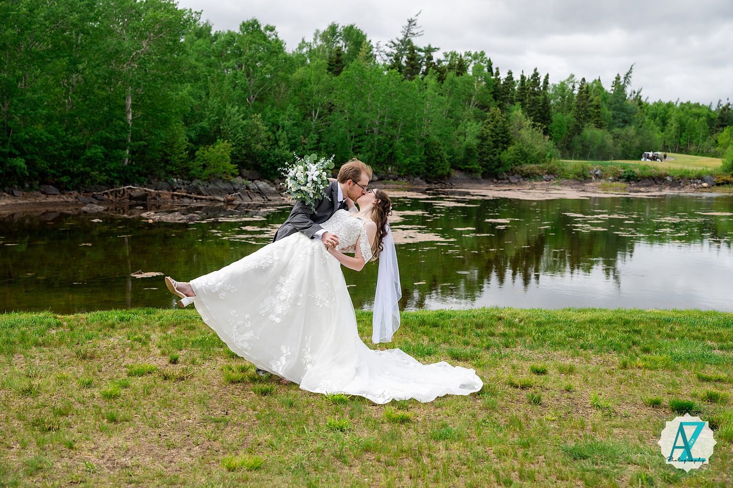 A romantic couple embraces near a serene pond surrounded by lush green trees on a cloudy day.