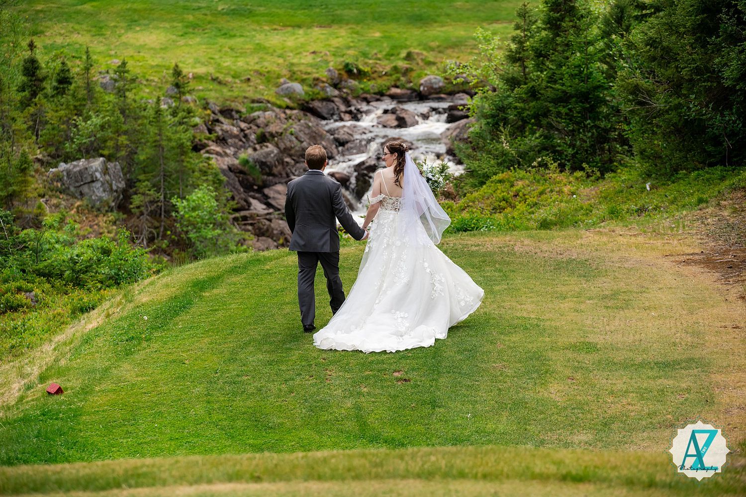 Two people in wedding attire walk hand in hand on a green path beside a rushing mountain stream.