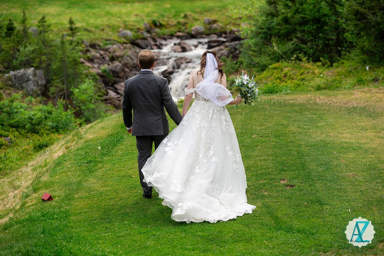A couple walks hand in hand across a lush green meadow with flowing white wedding attire in a scenic outdoor setting.