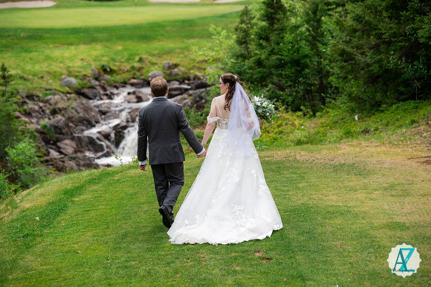 A couple walks along a grassy path next to a cascading waterfall on their wedding day at a scenic outdoor venue.