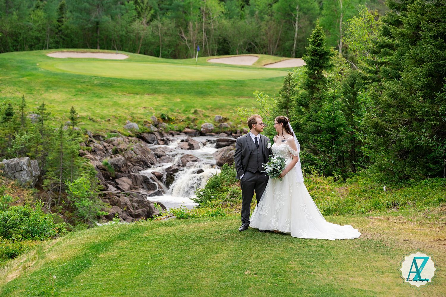 Two figures in wedding attire stand near a cascading waterfall on a scenic golf course with lush greenery.