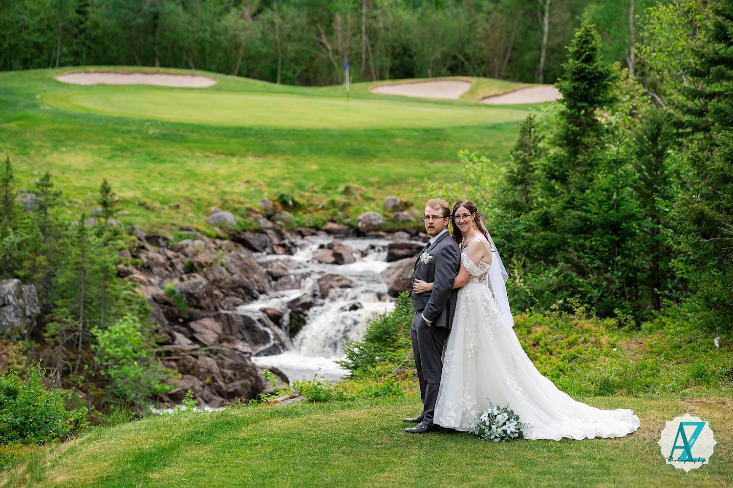 Couple poses on a golf course near a scenic rocky waterfall surrounded by evergreen trees and manicured greens.