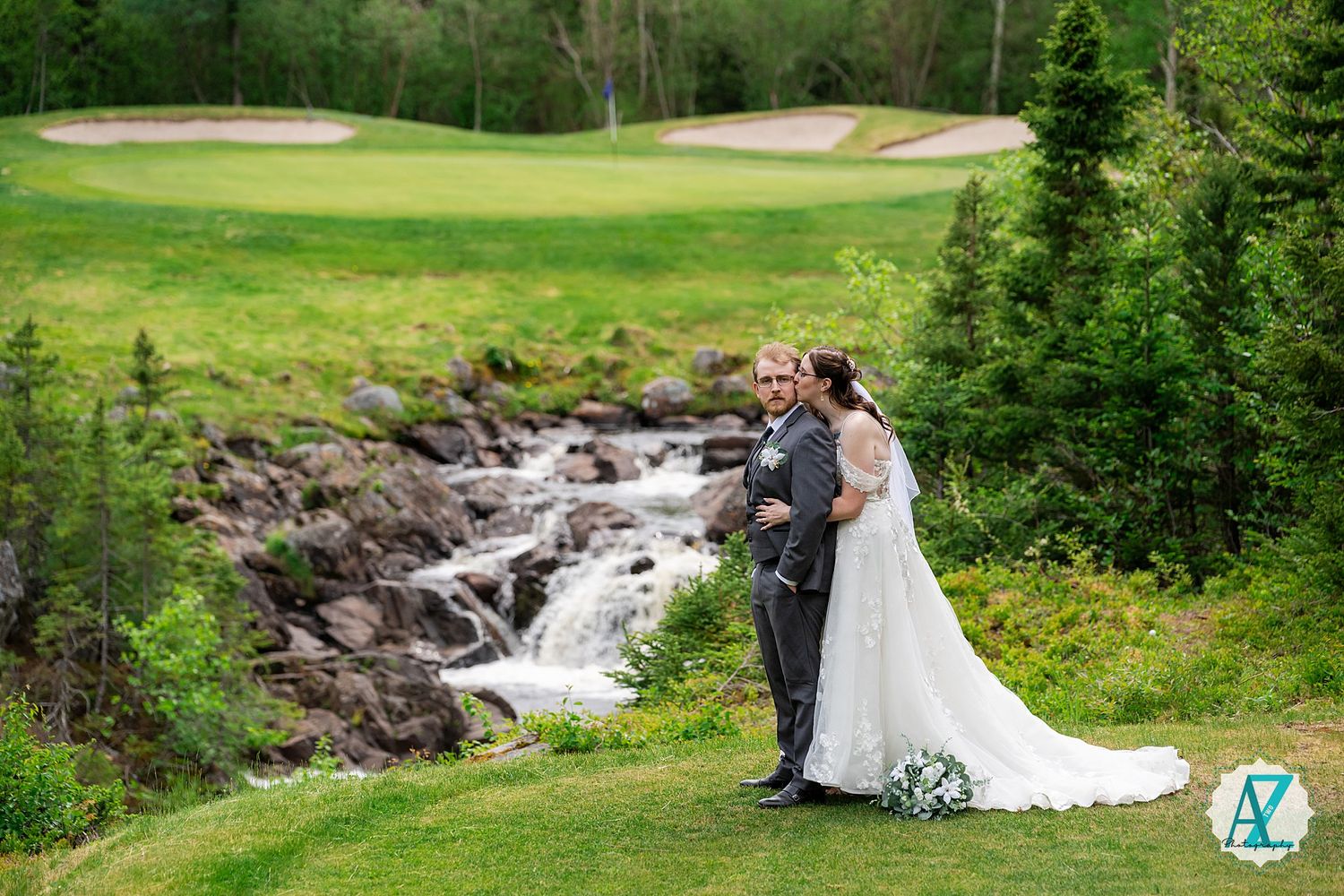 A couple in wedding attire embraces near a cascading waterfall on a golf course with sand bunkers visible in background.
