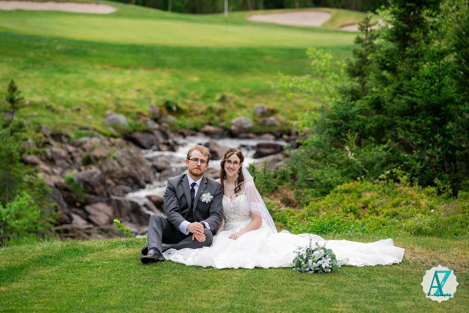 A wedding couple sits on grass beside a rocky stream at a golf course on their special day.