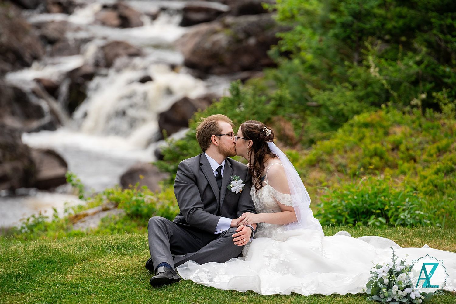 A couple in wedding attire sits on lush grass beside a scenic cascading waterfall in a natural outdoor setting.