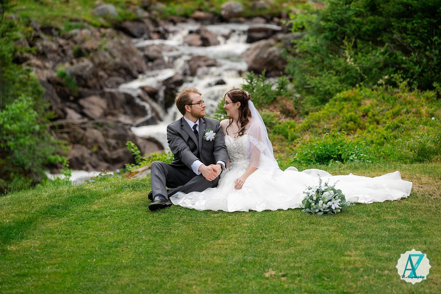 Couple in wedding attire sits on grass by cascading waterfall in scenic outdoor setting.