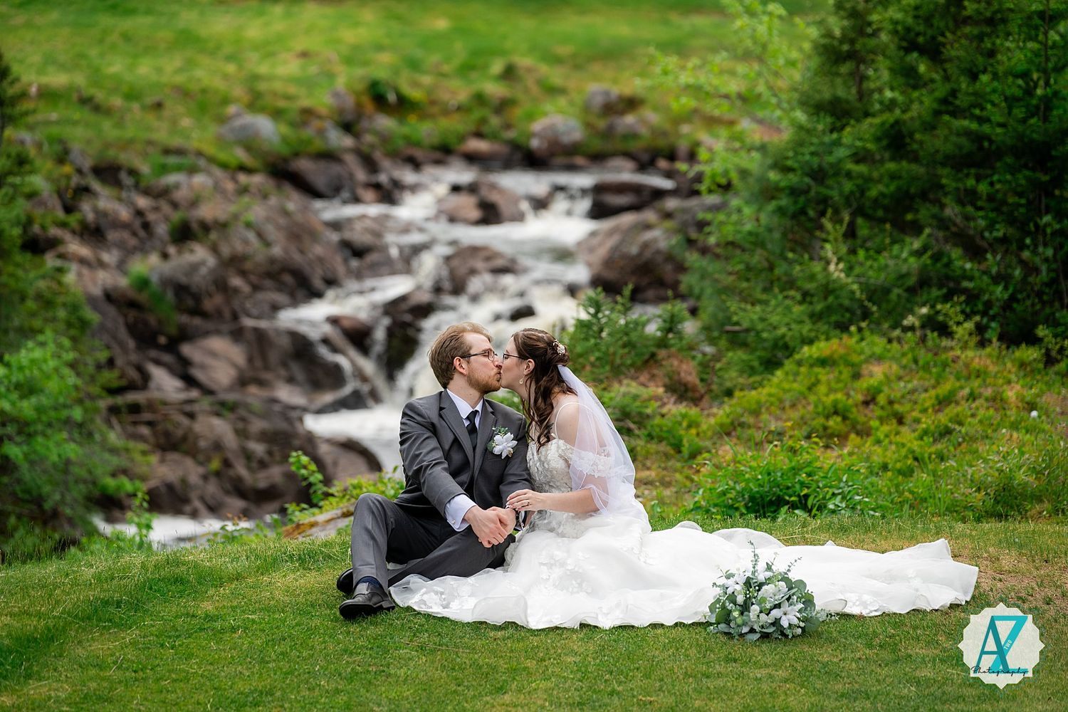 A couple sits intimately by a cascading mountain stream with lush greenery and rocks in the background.