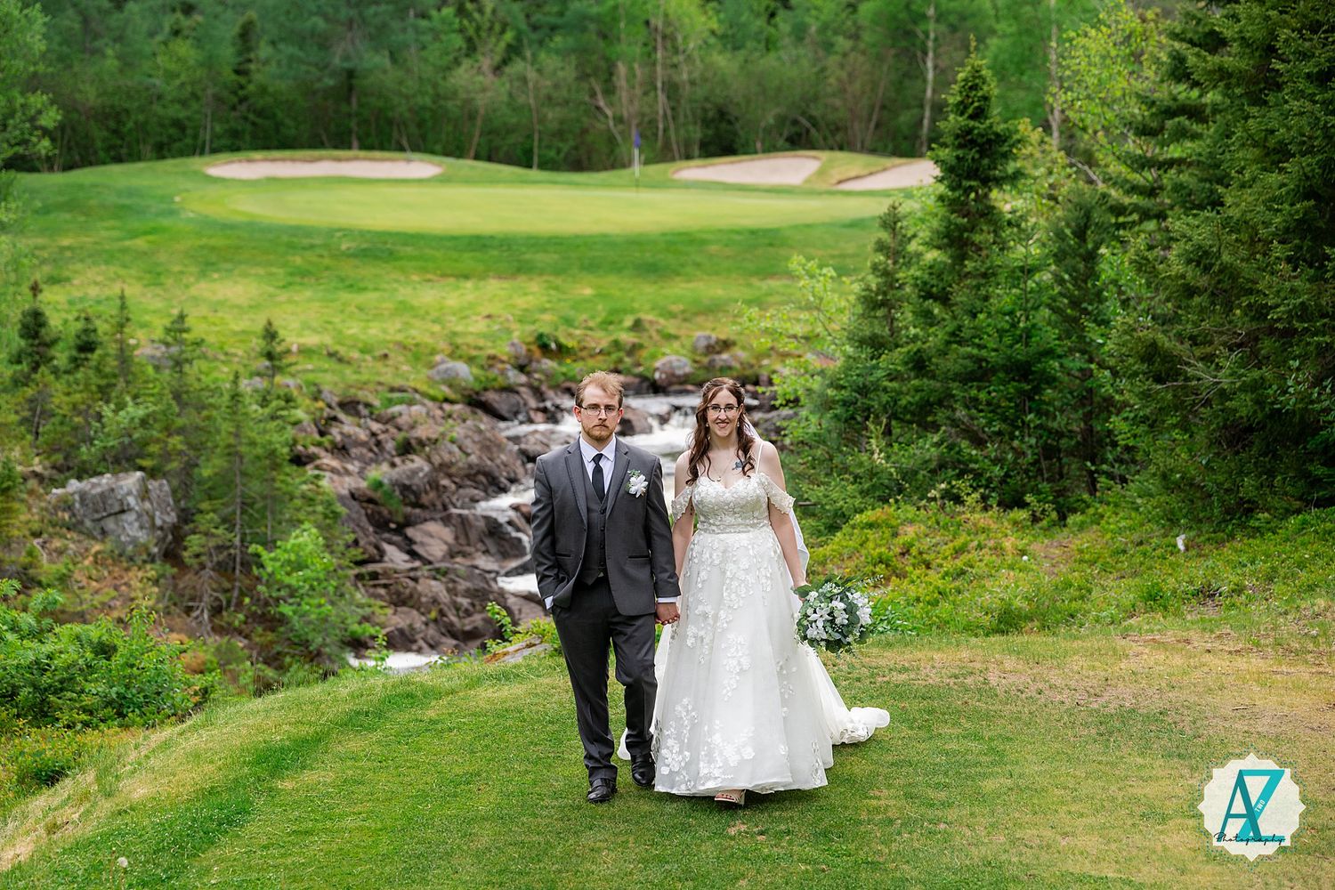 Two people walk together on a grassy path at a golf course with a waterfall and evergreen trees in the background.