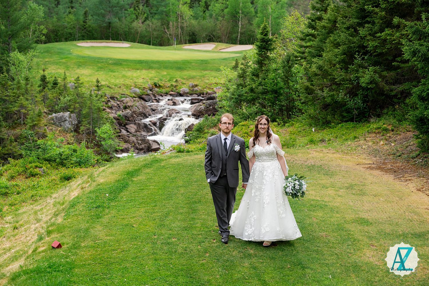 Wedding couple walking along a scenic golf course path with cascading waterfall and evergreen trees in the background.