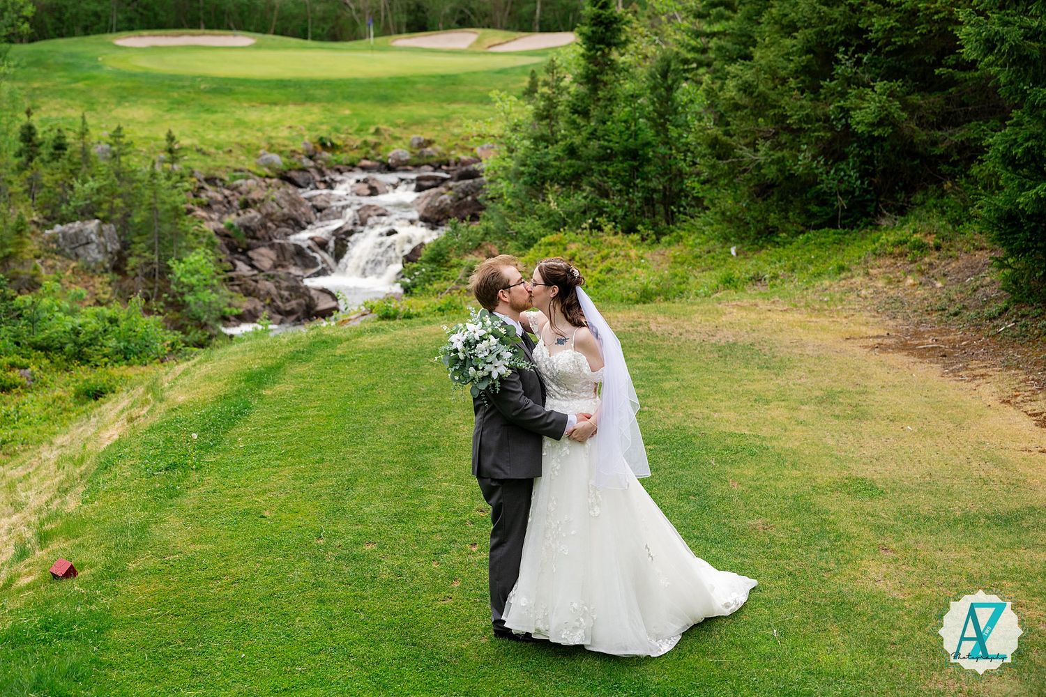 Two people in formal wedding attire embrace on a manicured golf course with a cascading waterfall in the background.