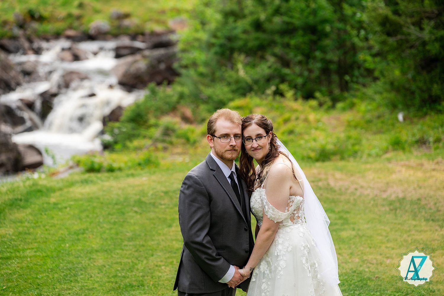 A couple in wedding attire poses together in front of a cascading waterfall and lush greenery.