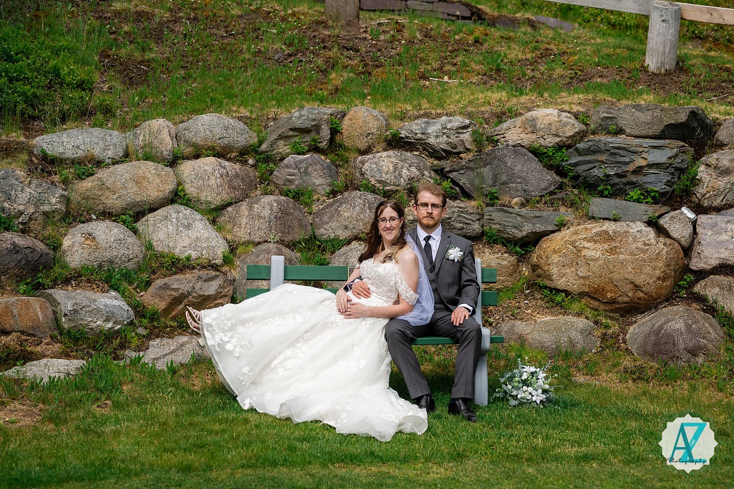 A couple sits together on a bench against a rustic stone wall backdrop surrounded by green grass.