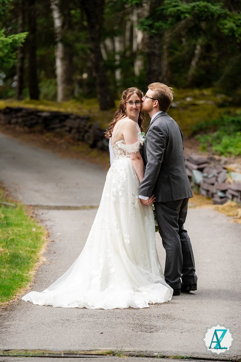 A couple shares an intimate moment on a paved forest path while dressed in elegant wedding attire.