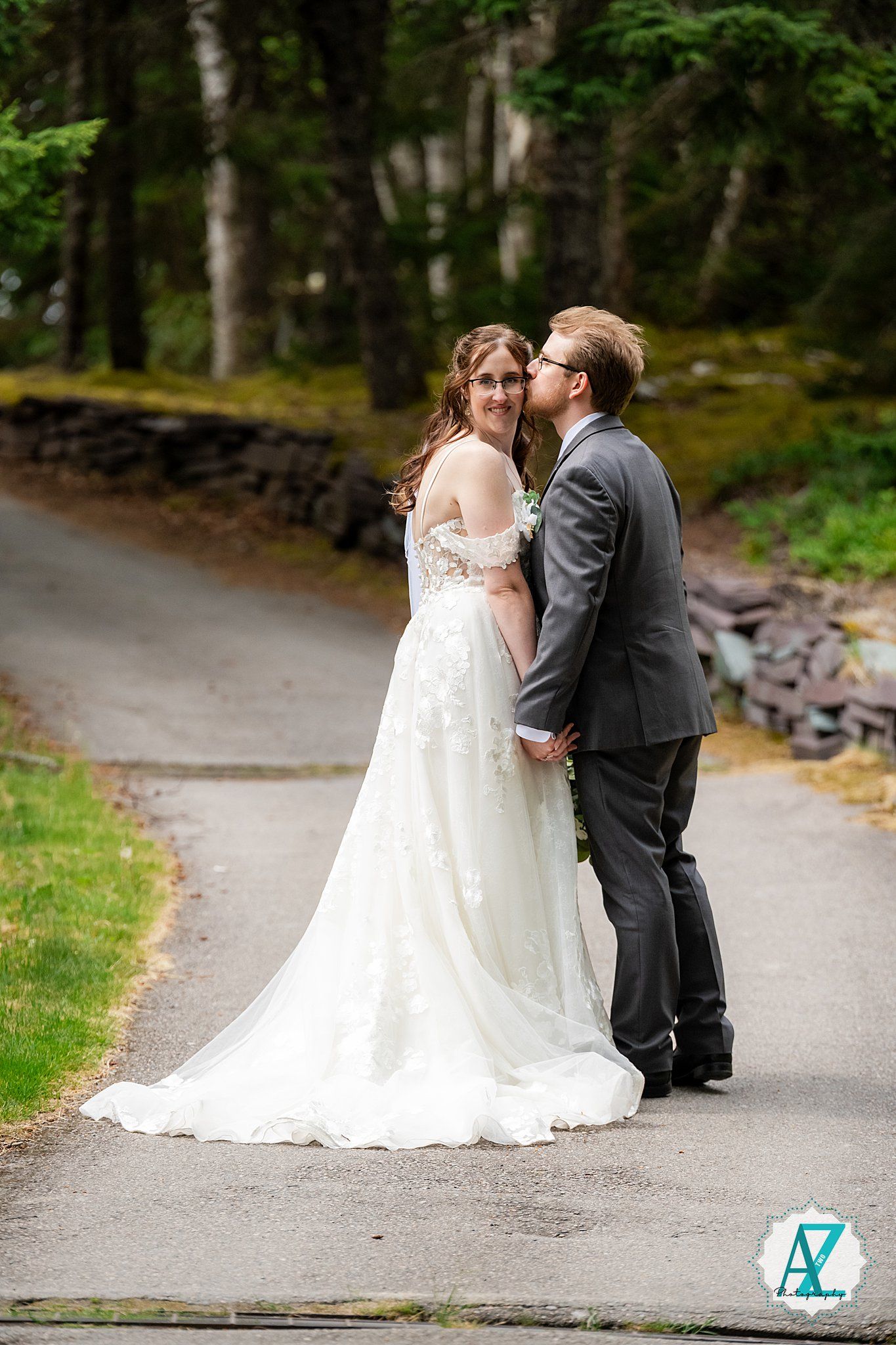 A couple shares an intimate moment on a paved forest path while dressed in elegant wedding attire.
