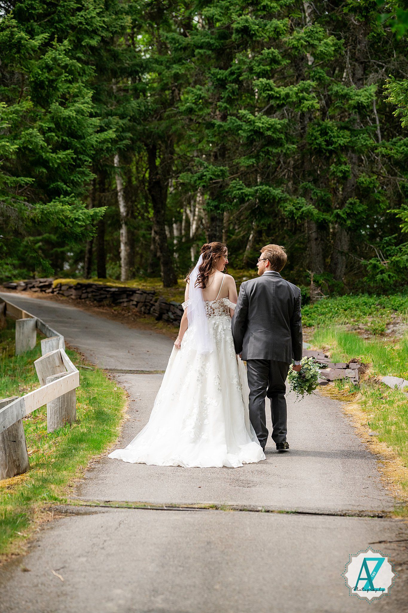 A couple walking down a serene tree-lined path with the bride wearing a flowing white gown and holding a bouquet.